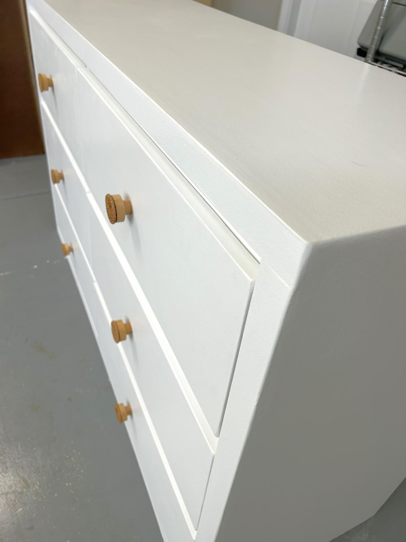 May include: A white dresser with four drawers and light wood knobs. The dresser is painted white and has a simple, modern design. The knobs are round and made of a light-colored wood.