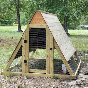May include: A-frame chicken coop constructed from light-coloured wood with a sloped roof. The coop has wire mesh sides and a small door with a latch. A water dispenser is visible inside. The coop is situated in a grassy outdoor area.
