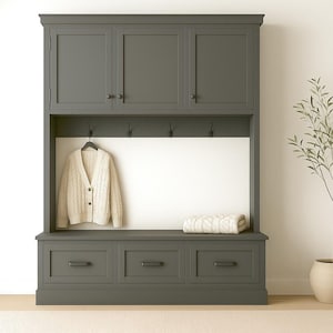 May include: A dark gray entryway storage unit with three drawers, upper cabinets, and a bench. A cream-colored cardigan hangs on a hook, and a folded blanket rests on the bench. A potted olive tree is to the right.