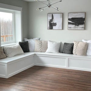 Puede incluir: Un banco blanco empotrado con una ventana y una colección de almohadas. Las almohadas son de varios colores y texturas, incluyendo beige, gris y blanco. Dos obras de arte abstractas cuelgan en la pared sobre el banco.