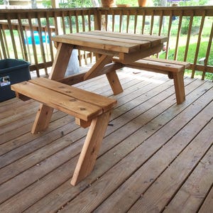 Op de afbeelding: Een houten picknicktafelset met aangehechte banken. De tafel en banken zijn gemaakt van lichtgekleurd hout en hebben een klassiek A-frame ontwerp. De set staat buiten op een houten terras.