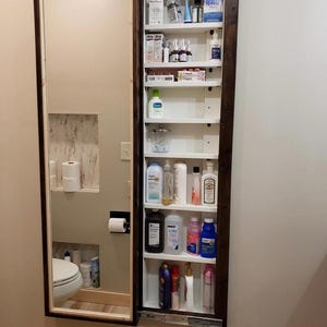 Può includere: Un mobiletto a specchio con cornice scura, montato su una parete chiara. L'armadietto è aperto, rivelando ripiani pieni di vari articoli da toeletta, farmaci e articoli per la cura della persona. Lo specchio riflette un water e altri sanitari.