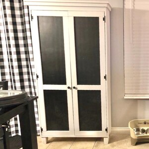 May include: A white cabinet with two doors and black panels. The cabinet has a rectangular shape and is standing on a tiled floor. The cabinet doors have black handles. The cabinet is next to a black and white checkered curtain.