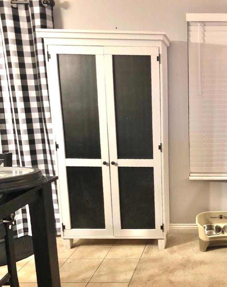 May include: A white cabinet with two doors and black panels. The cabinet has a rectangular shape and is standing on a tiled floor. The cabinet doors have black handles. The cabinet is next to a black and white checkered curtain.