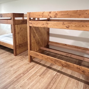 May include: Two wooden bunk beds with a natural finish. The beds are made of thick planks and have a simple, sturdy design. One bed has a mattress, while the other is empty. The beds are in a room with wood flooring.