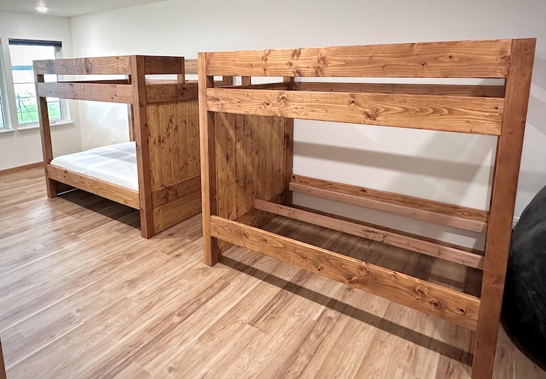 May include: Two wooden bunk beds with a natural finish. The beds are made of thick planks and have a simple, sturdy design. One bed has a mattress, while the other is empty. The beds are in a room with wood flooring.