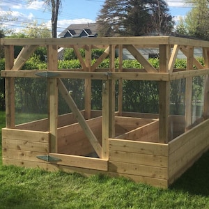 May include: A wooden raised garden bed with a mesh and clear panel enclosure. The structure is made of light-colored wood and features a slatted design. The garden bed is set on a grassy lawn, with a house and trees in the background.