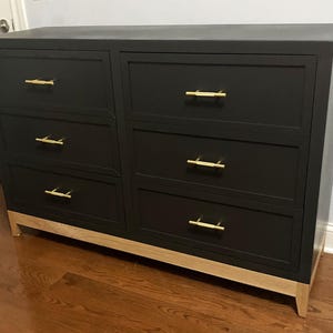 May include: A black six-drawer dresser with gold-colored handles and light wood-colored legs. The dresser is rectangular and has a modern design, suitable for bedroom storage.