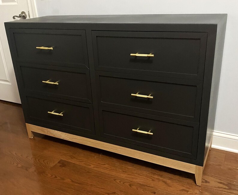 May include: A black six-drawer dresser with gold-colored handles and light wood-colored legs. The dresser is rectangular and has a modern design, suitable for bedroom storage.