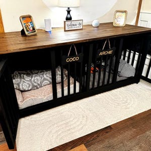 Puede incluir: Una jaula negra para perros con una parte superior de madera, con dos compartimentos con los nombres "Coco" y "Archie" en etiquetas con forma de hueso. La jaula incluye ropa de cama y se coloca sobre una alfombra de color claro. Una foto enmarcada y un letrero que dice "welcome-ish" están en la parte superior.