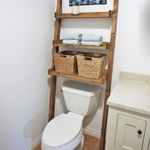 Op de afbeelding: Een houten ladderplank in een badkamer. De plank heeft drie planken met opgevouwen handdoeken en geweven manden. Een wit toilet staat voor de plank. Een decoratief stuk hangt aan de muur.