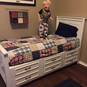 May include: A white bed frame with six drawers, a patchwork quilt, and a blue pillow. A young child in pajamas is jumping on the bed. A framed artwork and a small wall-mounted storage unit are visible in the background.
