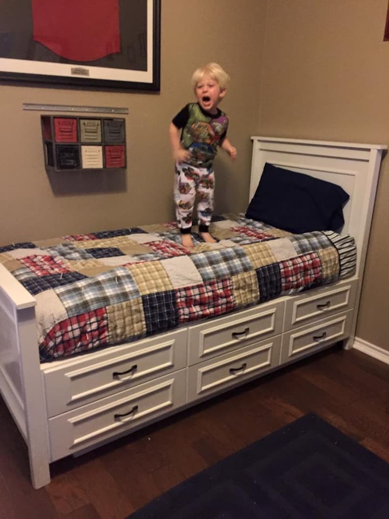 May include: A white bed frame with six drawers, a patchwork quilt, and a blue pillow. A young child in pajamas is jumping on the bed. A framed artwork and a small wall-mounted storage unit are visible in the background.