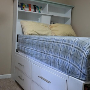 May include: A white bed frame with built-in drawers and a headboard with shelves. The bed is covered with a blue and gray plaid comforter and two yellow pillows. The headboard has the word "HOLLY" on it.