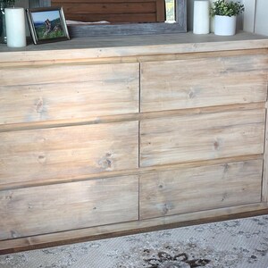 May include: A six-drawer wooden dresser with a light, natural finish. The dresser has a rectangular shape with six equally sized drawers. The top of the dresser has decorative items, including a framed picture, candles, and a small potted plant.