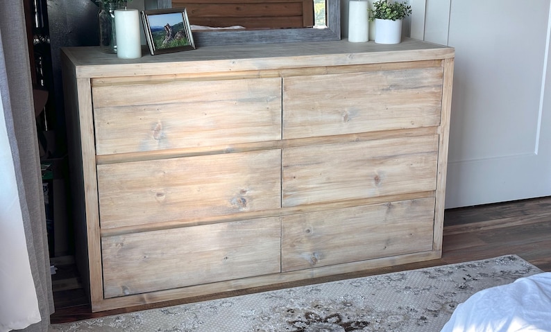 May include: A six-drawer wooden dresser with a light, natural finish. The dresser has a rectangular shape with six equally sized drawers. The top of the dresser has decorative items, including a framed picture, candles, and a small potted plant.