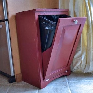 Puede incluir: Un cubo de basura de madera roja con una puerta con bisagras y un tirador en forma de corazón. La puerta está entreabierta, revelando una bolsa de basura negra en el interior. El cubo de basura está colocado junto a un frigorífico y un armario.