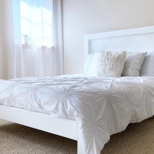 May include: A white bed frame with a matching headboard. The bed is made with a white comforter and pillows. The room has a neutral color palette with a window and a piece of art on the wall.