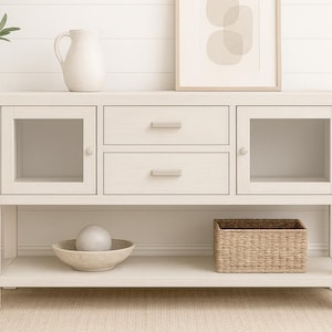 以下が含まれることがあります： 2つの引き出しと2つのガラス扉キャビネットを備えた、明るい色の木製コンソールテーブル。装飾的なピッチャー、アートワーク、織りバスケットがテーブルにあります。鉢植えの植物と装飾的なボウルが近くにあります。