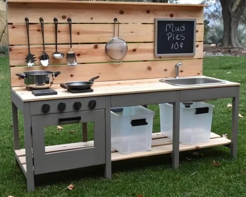 May include: A wooden outdoor play kitchen with a gray frame, featuring a sink, stove, and storage. It includes a chalkboard sign that reads "Mud Pies 10$" and hanging cooking utensils. The kitchen is set on a grassy lawn.