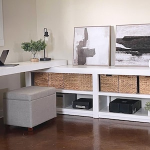 May include: A modern white desk and storage unit with woven baskets, abstract art, and a gray ottoman. A laptop and lamp sit on the desk. The room is decorated in neutral tones, creating a minimalist aesthetic.