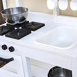 May include: A white wooden play kitchen with a stainless steel bowl on black stovetop burners. The kitchen features a white sink, black knobs, and a handle. Wooden spoons hang above the countertop.