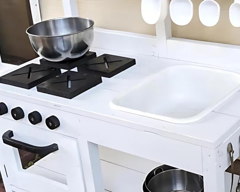 May include: A white wooden play kitchen with a stainless steel bowl on black stovetop burners. The kitchen features a white sink, black knobs, and a handle. Wooden spoons hang above the countertop.