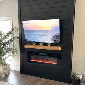 Peut inclure: Un meuble de divertissement mural noir avec une cheminée électrique intégrée et une télévision affichant un coucher de soleil sur la mer. Une étagère en bois contient de petites plantes en pot. L'ensemble est placé contre un mur neutre.