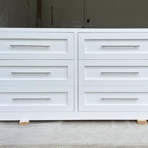 May include: A white six-drawer dresser with silver-toned handles. The dresser is rectangular and sits on small wooden blocks. The drawers are evenly spaced and have a simple, modern design.