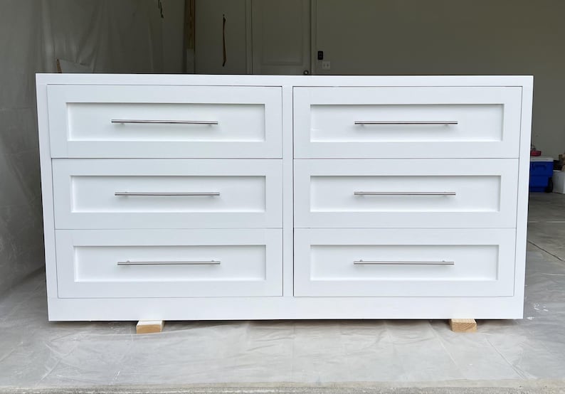 May include: A white six-drawer dresser with silver-toned handles. The dresser is rectangular and sits on small wooden blocks. The drawers are evenly spaced and have a simple, modern design.