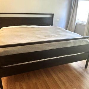 May include: A dark brown wooden bed frame with a headboard and footboard. The bed is covered with a light-colored blanket. The bed frame has a simple, modern design and is set on a hardwood floor.