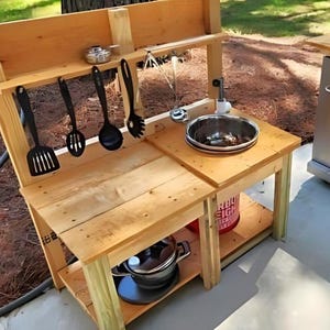 May include: A wooden outdoor play kitchen with a sink, shelves, and hanging utensils. The kitchen has a natural wood finish and includes a stainless steel sink, pots, and a red bucket. Black cooking utensils hang above the countertop.