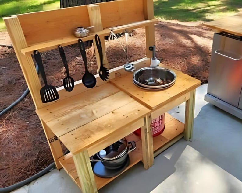 May include: A wooden outdoor play kitchen with a sink, shelves, and hanging utensils. The kitchen has a natural wood finish and includes a stainless steel sink, pots, and a red bucket. Black cooking utensils hang above the countertop.