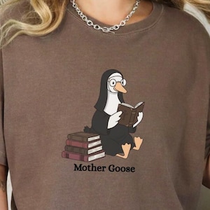 May include: A brown t-shirt featuring a cartoon nun reading a book, with the text "Mother Goose" below. The shirt is shown in the color Espresso. The brand name "Pax Sanctorum" is at the bottom of the image.