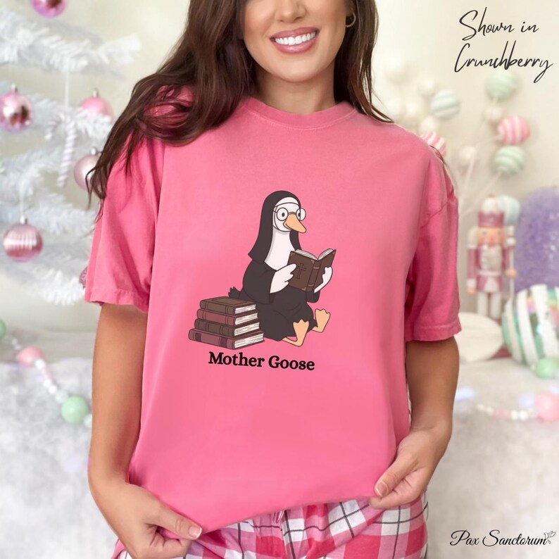 K&ouml;nnte beinhalten: Ein pinkfarbenes T-Shirt mit einer Cartoon-Gans, die als Nonne verkleidet ist und ein Buch liest. Die Gans sitzt auf einem Stapel B&uuml;cher, darunter steht "Mother Goose". Das Shirt hat eine einfarbige pinke Farbe.