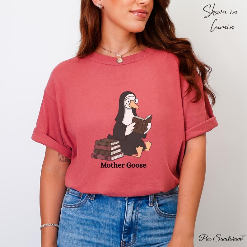 K&ouml;nnte beinhalten: Ziegelrotes T-Shirt mit einer Cartoon-Gans, die als Nonne verkleidet ist und ein Buch liest. Die Gans sitzt auf einem Stapel B&uuml;cher, darunter steht "Mother Goose". Das Shirt wird in Cumin gezeigt.