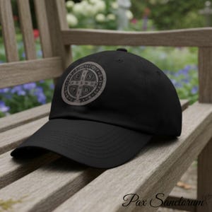 Peut inclure: Casquette de baseball noire ornée d'un écusson circulaire avec des symboles et du texte religieux de couleur marron clair. La casquette a une visière incurvée et une couronne structurée. Le texte "Pax Sanctorum" est imprimé en cursive en bas.