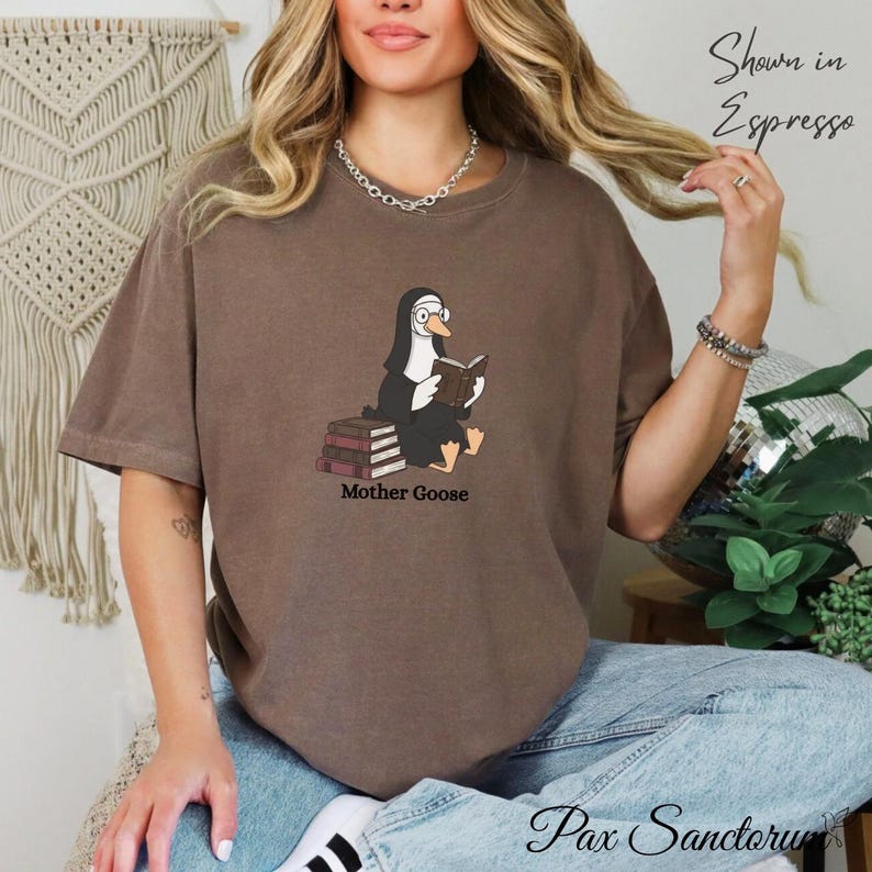 May include: A brown t-shirt featuring a cartoon nun reading a book, with the text "Mother Goose" below. The shirt is shown in the color Espresso. The brand name "Pax Sanctorum" is at the bottom of the image.