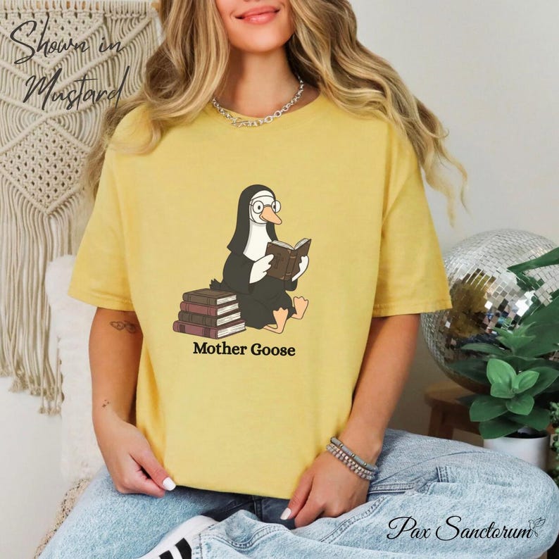 K&ouml;nnte beinhalten: Senffarbenes T-Shirt mit einer Cartoon-Gans in Nonnenkluft, die ein Buch liest. Die Gans sitzt auf B&uuml;chern, darunter steht "Mother Goose". Das Shirt hat eine lockere Passform. Der Text "Shown on Mustard" ist ebenfalls sichtbar.