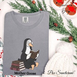 K&ouml;nnte beinhalten: Ein graues Comfort Colors XL-TG-EG T-Shirt mit einer Cartoon-Gans in Nonnentracht, die eine Brille tr&auml;gt und ein Buch liest. Der Text "Mother Goose" ist darunter gedruckt. Das Shirt ist Teil einer festlichen Kollektion.