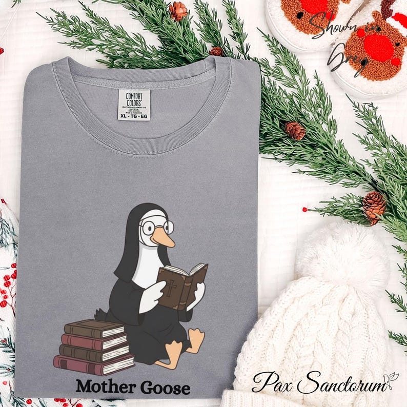 K&ouml;nnte beinhalten: Ein graues Comfort Colors XL-TG-EG T-Shirt mit einer Cartoon-Gans in Nonnentracht, die eine Brille tr&auml;gt und ein Buch liest. Der Text "Mother Goose" ist darunter gedruckt. Das Shirt ist Teil einer festlichen Kollektion.