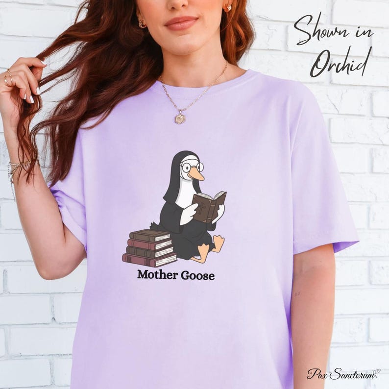 K&ouml;nnte beinhalten: Helllila T-Shirt mit einer Cartoon-Gans in Nonnentracht, die ein Buch liest und eine Brille tr&auml;gt. Der Text "Mother Goose" ist darunter gedruckt. Das T-Shirt ist mit "Shown in Orchid" gekennzeichnet.