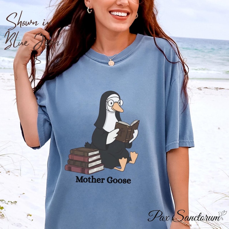K&ouml;nnte beinhalten: Hellblaues T-Shirt mit einer Cartoon-Gans in Nonnenkluft, die ein Buch liest. Unter der Illustration steht der Text "Mother Goose". Das Shirt tr&auml;gt auch die Aufschrift "Shown in Blue" und "Pax Sanctorum".