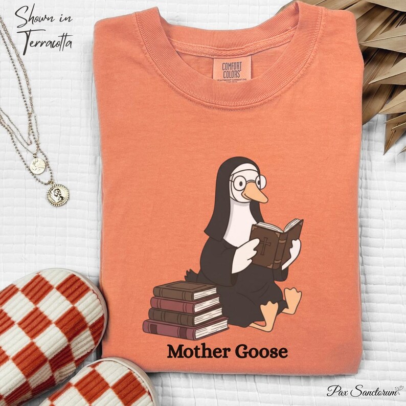 K&ouml;nnte beinhalten: Terracottafarbenes T-Shirt mit einer Cartoon-Gans, die als Nonne gekleidet ist, eine Brille tr&auml;gt und ein Buch liest. Der Text "Mother Goose" ist darunter gedruckt. Das Shirt ist gefaltet und auf einer wei&szlig;en Oberfl&auml;che ausgestellt.