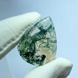 May include: A teardrop-shaped gemstone with a clear base and intricate green and white inclusions, resembling a landscape. The stone is held by a gloved hand, showcasing its polished surface and natural patterns. The gem's unique design makes it a striking piece.