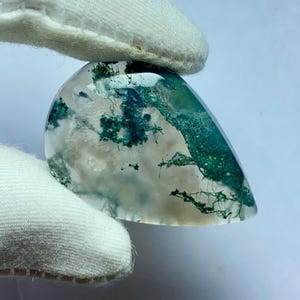 May include: A polished, teardrop-shaped gemstone with a translucent base and intricate green moss-like inclusions. The stone is held by a gloved hand, showcasing its natural beauty and unique patterns. The gemstone's surface is smooth and reflective.
