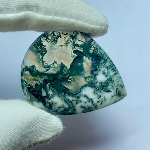 May include: A teardrop-shaped moss agate gemstone. The stone features a mix of green, white, and tan colors, with intricate patterns resembling moss. The gemstone is held by a gloved hand, showcasing its polished surface.
