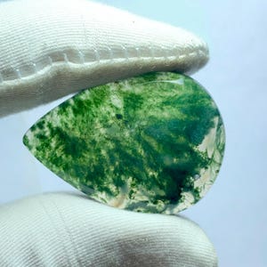 May include: A teardrop-shaped moss agate gemstone, held between gloved fingers. The translucent stone features vibrant green, fern-like inclusions, creating a natural, artistic pattern. The stone's polished surface reflects light, showcasing its unique beauty.