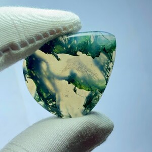 May include: A translucent, heart-shaped gemstone with green, moss-like inclusions. The stone is held by a gloved hand, showcasing its natural patterns and colors. The gemstone has a polished surface and a unique, organic appearance.