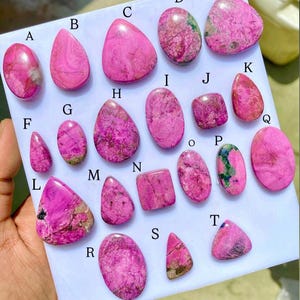 May include: A collection of polished, vibrant pink gemstones in various shapes, including oval, teardrop, and rectangular. Some stones display darker veining and patches of green. The gemstones are arranged on a white surface, with letters labeling each stone.
